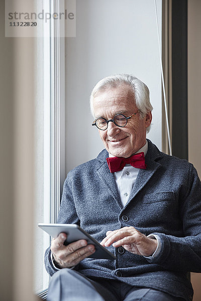 Lächelnder älterer Mann auf der Fensterbank sitzend mit Tablette