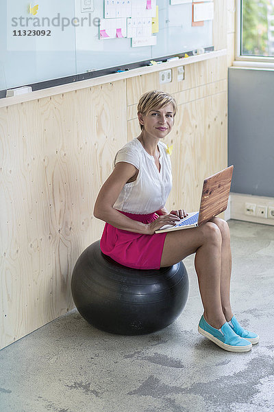 Lächelnde Geschäftsfrau sitzt auf Fitnessball mit Laptop