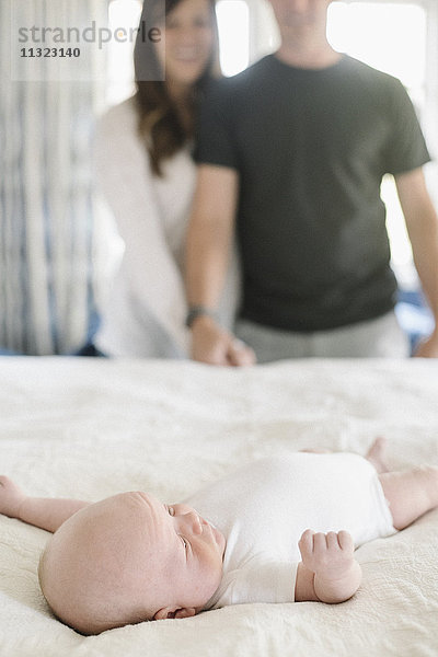 Zwei Eltern halten sich an den Händen und stehen über einem kleinen Baby  das auf einem Bett liegt.