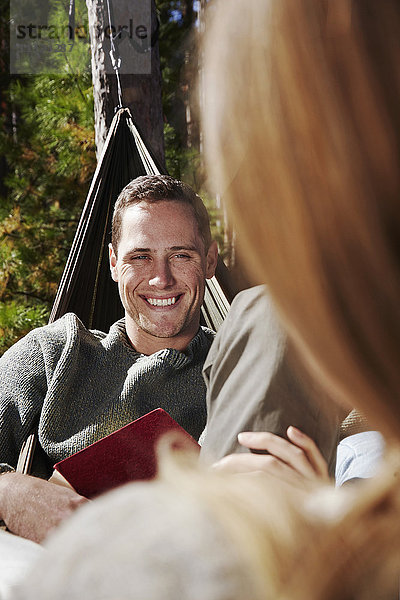 Zwei Personen sitzen in Hängematten im Freien. Ein Mann  der einen Begleiter anlächelt.