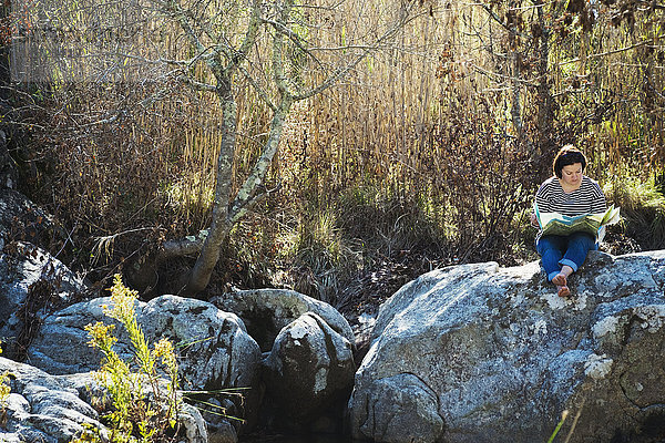 Eine Frau sitzt auf den Felsen im Schatten eines Flusses und schaut auf eine Karte.
