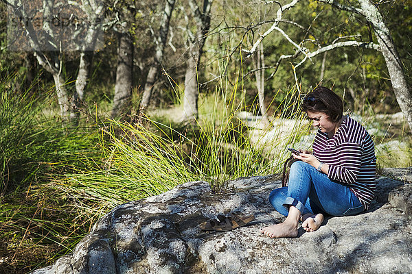 Eine Frau sitzt auf den Felsen im Schatten eines Flusses und schaut auf ein Smartphone.
