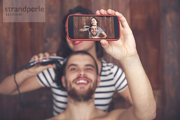 Junger Mann  der einen Selfie nimmt  während er sich von seiner Freundin mit einer Haarschneidemaschine die Haare schneiden lässt.