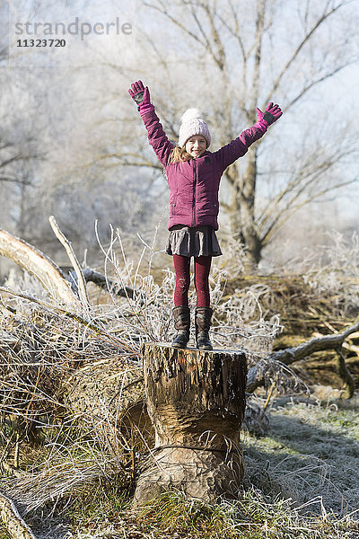 Porträt eines glücklichen Mädchens mit erhobenen Armen auf Baumstamm im Winter