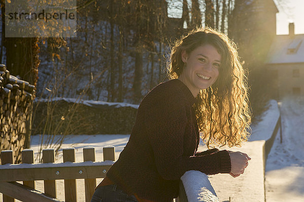Porträt einer glücklichen jungen Frau  die sich im Gegenlicht auf ein schneebedecktes Geländer stützt.