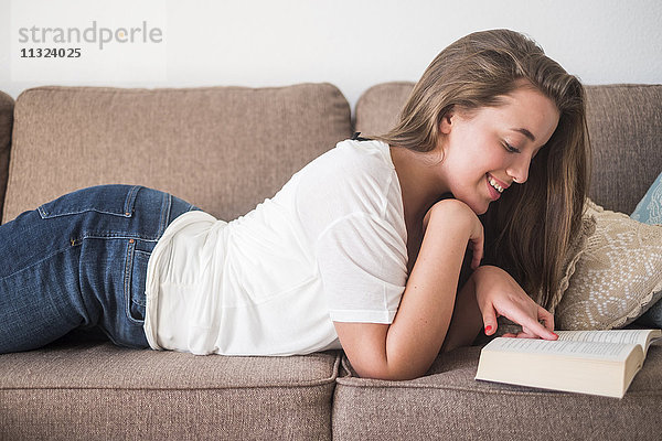 Lächelnde junge Frau auf der Couch liegend Lesebuch