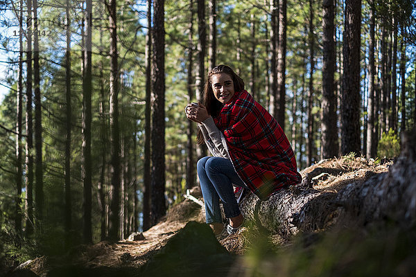Lächelnde junge Frau mit Decke und Getränk auf Felsen im Wald sitzend