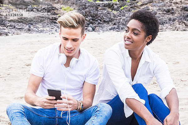 Junger Mann sitzend neben Freundin am Strand mit Handy und Kopfhörer