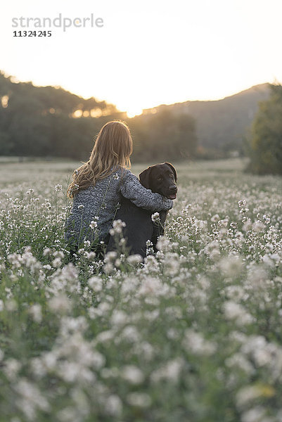 Junge Frau und ihr Hund sitzen in der Dämmerung im Blumenfeld.