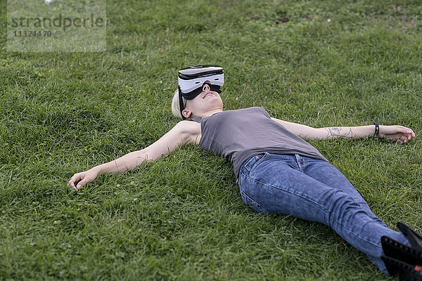 Junge Frau liegt auf einer Wiese und trägt eine Virtual Reality Brille im Freien.