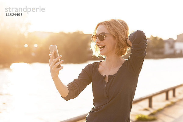 Fröhliche junge Frau nimmt Selfie mit Smartphone im Gegenlicht