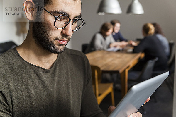 Junger Mann mit digitalem Tablett im modernen Büro mit Mitarbeitern am Tisch im Hintergrund