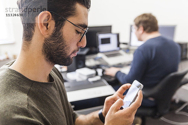 Junger Mann mit Smartphone im modernen Büro mit Mitarbeiter im Hintergrund