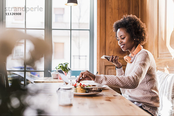 Lächelnde junge Frau mit Handy und Tablette im Cafe