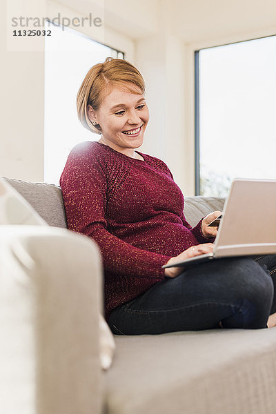 Lächelnde schwangere Frau auf der Couch online einkaufen