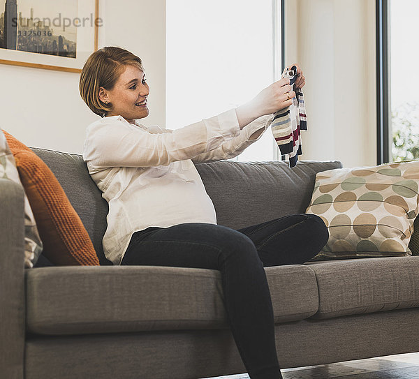 Schwangere Frau auf der Couch mit Babykleidung
