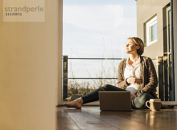 Schwangere Frau sitzt auf dem Boden mit Laptop und genießt die Sonne.