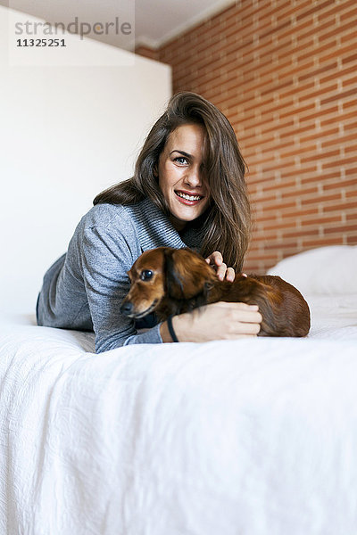 Lächelnde junge Frau mit ihrem Hund im Bett liegend