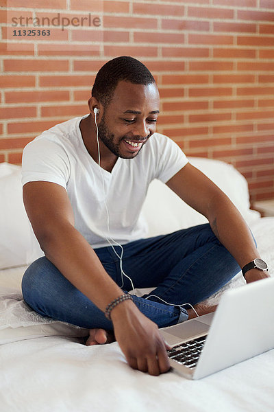 Lächelnder junger Mann mit Kopfhörern auf dem Bett sitzend mit Laptop