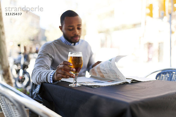 Junger Mann liest Zeitung und trinkt ein Bier im Straßencafé.