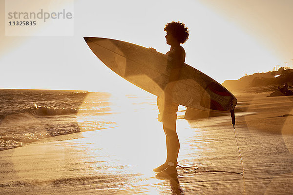 Junger Mann mit Surfbrett am Strand bei Sonnenuntergang