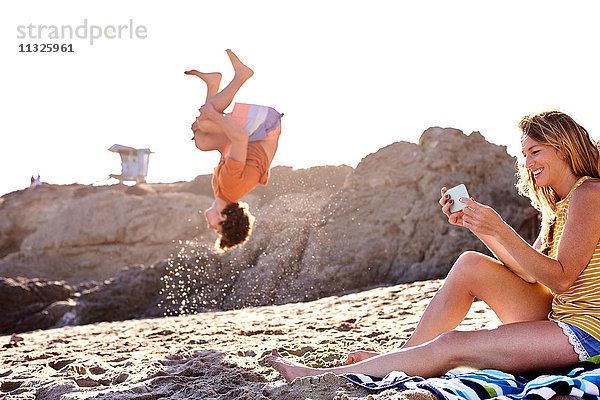 Junger Mann am Strand beim Salto mit Frau beim Handy-Checken