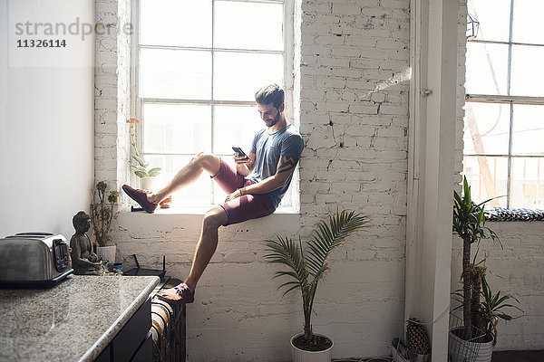 Junger Mann sitzt auf der Fensterbank und schaut auf ein Handy in einem Loft.