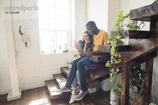 Lächelndes junges Paar sitzt auf einer Treppe in einem Loft und teilt sich ein Handy.