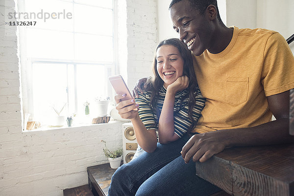 Ein glückliches junges Paar sitzt auf einer Treppe in einem Loft und teilt sich ein Handy.