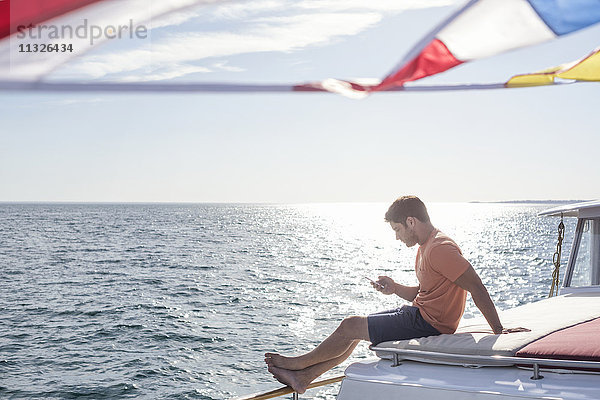 Junger Mann sitzt auf einem Boot und schaut aufs Handy.