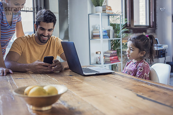 Familie in der Küche sitzend  Eltern mit Smartphone  Tochter mit Blick auf Laptop