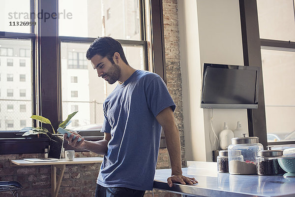 Junger Mann steht in der Küche mit Smartphone