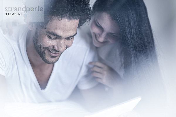 Lächelndes junges Paar im Bett mit Blick auf Tablette