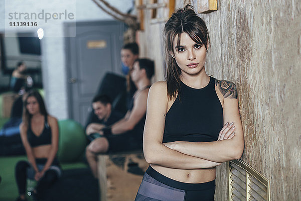 Porträt einer selbstbewussten Athletin mit gekreuzten Armen und Freunden im Hintergrund im Fitnessstudio.
