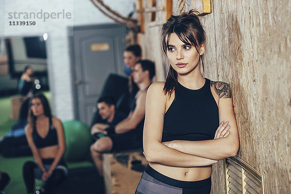 Nachdenkliche Sportlerin mit gekreuzten Armen und Freunden im Hintergrund im Fitnessstudio