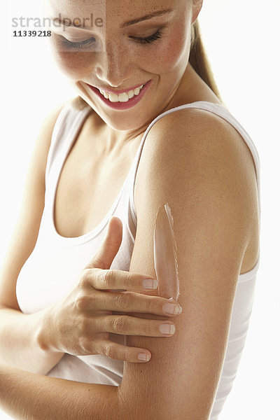 Fröhliche Frau beim Auftragen von Körperlotion auf den Arm vor weißem Hintergrund