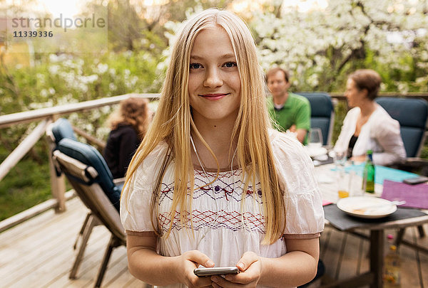 Porträt eines jungen Mädchens mit einem Smartphone im Hof und einer Familie im Hintergrund.