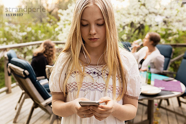 Teenager-Mädchen mit Smartphone im Hof mit Familie im Hintergrund