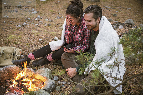 Hochwinkelansicht des glücklichen Paares in Decke gehüllt bei der Benutzung des Mobiltelefons durch die Feuerstelle auf dem Campingplatz.