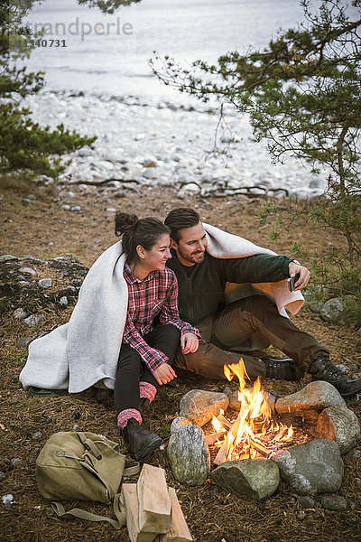 Paar in Decke gehüllt  das sich beim Sitzen an der Feuerstelle auf dem Campingplatz selbst mitnimmt