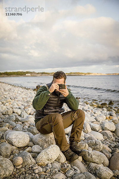 Mann  der von der Kamera fotografiert  während er auf einem Felsen am Strand gegen den Himmel sitzt.