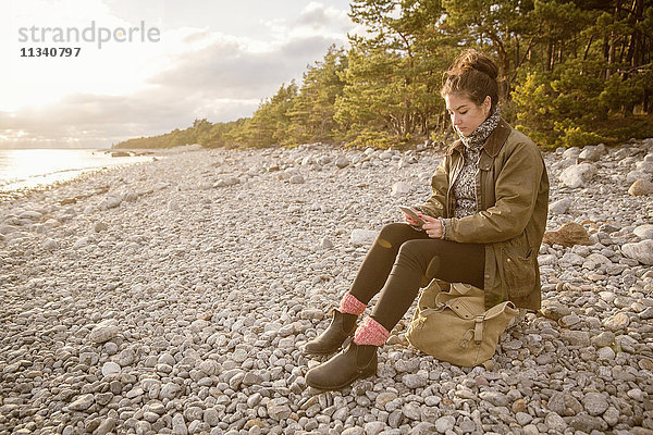 Frau sitzt auf einem Felsen mit Tasche  während sie am Strand mit dem Handy telefoniert.