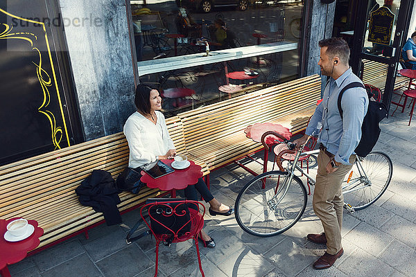 Geschäftsmann mit Fahrrad im Gespräch mit einer Kollegin  die im Straßencafé sitzt.