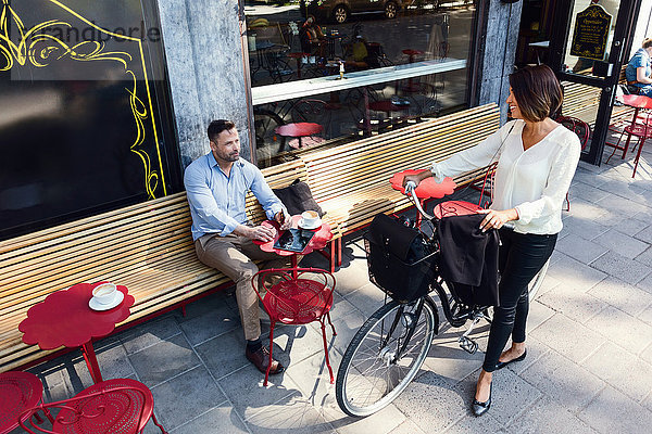 Geschäftsfrau mit Fahrrad im Gespräch mit einem männlichen Kollegen im Straßencafé
