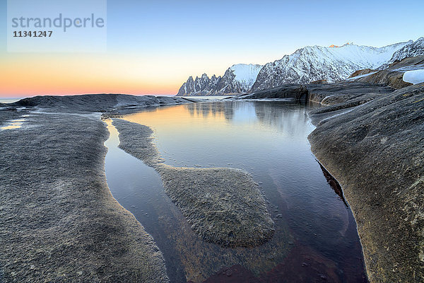 Orangefarbener Himmel bei Sonnenuntergang spiegelt sich auf schneebedeckten Gipfeln und dem gefrorenen Meer  das von Felsen umgeben ist Tungeneset  Senja  Troms County  Arktis  Norwegen  Skandinavien  Europa