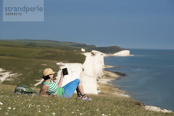 Eine Frau liest auf den Klippen mit Blick auf die Seven Sisters-Küste in der Ferne auf ihrem iPad  South Downs National Park  East Sussex  England  Vereinigtes Königreich  Europa