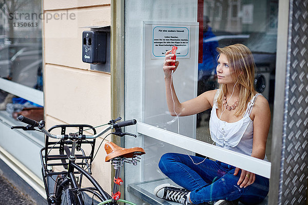 Junge Frau mit Handy und Ohrhörern hinter einer Fensterscheibe