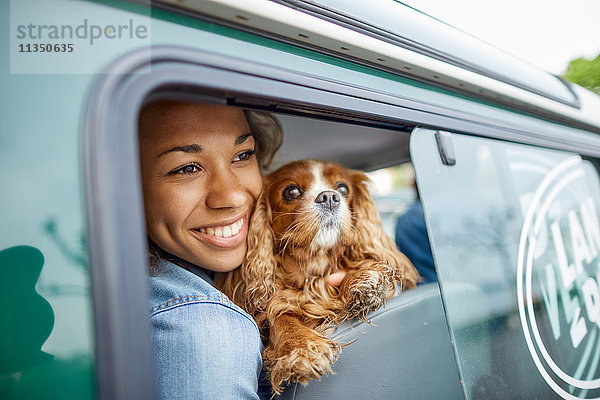 Fröhliche junge Frau mit Hund im Auto schaut aus dem Fenster