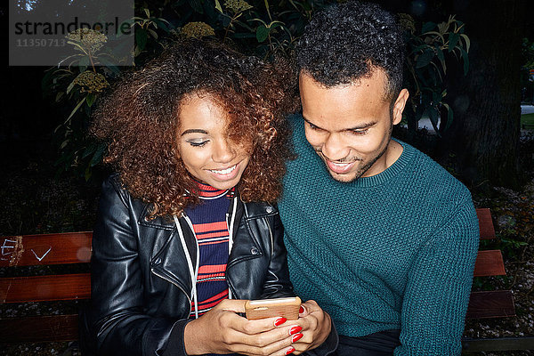 Lächelndes junges Paar auf einer Parkbank schaut aufs Handy