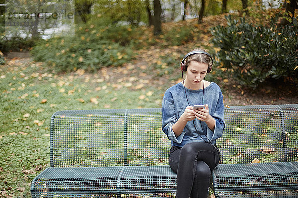 Junge Frau mit Kopfhörern auf einer Parkbank schaut auf ihr Handy